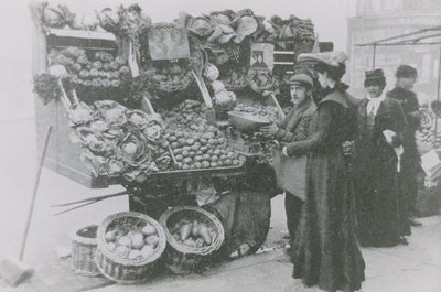 Un épicier à côté de son étal servant une femme - English Photographer